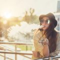 Pretty young hipster woman in black hat vape ecig, vaping device at the sunset. Toned image.